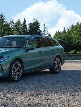 VW Der neue Passat VW Passat in blaugrün steht auf einer Kiesfläche, das Gelände grenzt an einen Tannenwald.